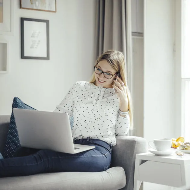 Young woman sat on a sofa using a phone with an open laptop